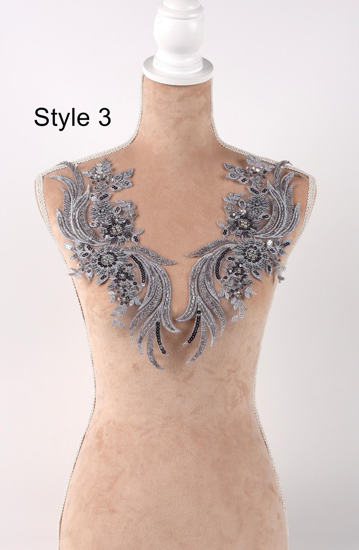A mannequin wearing a beige dress with intricate silver and black lace appliques on the shoulders.