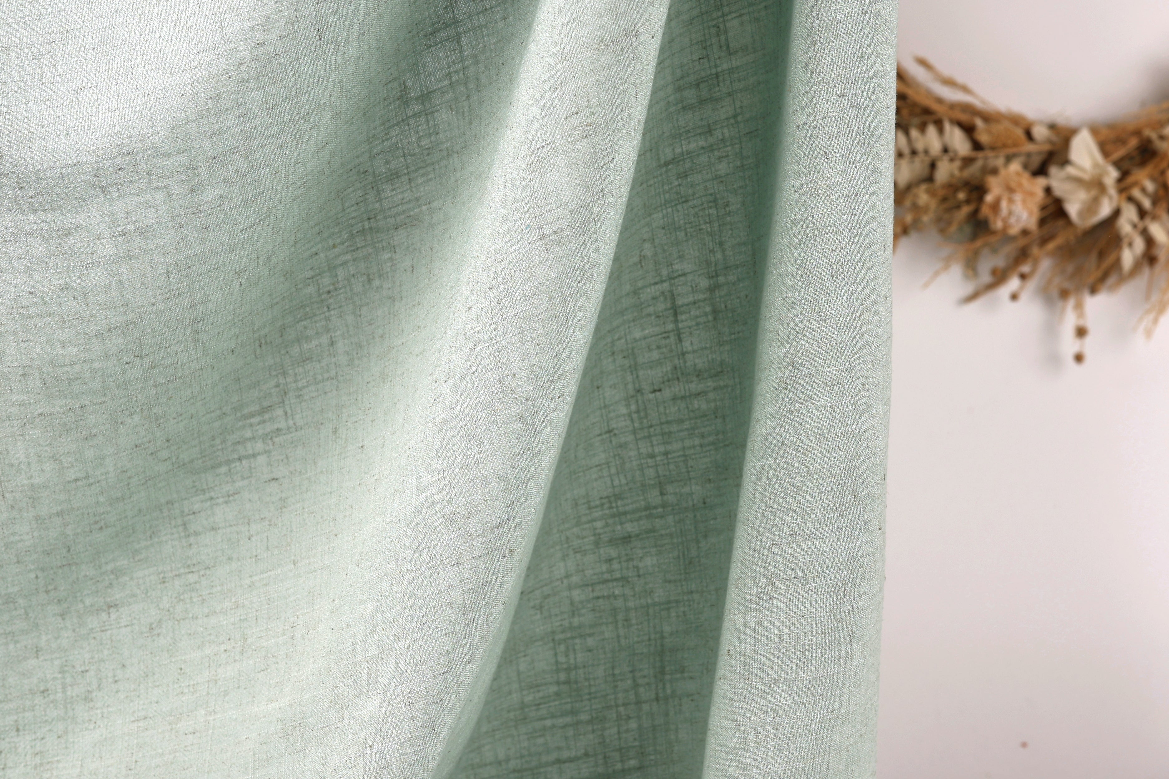 a close-up view of a light green fabric, possibly a curtain or textile, with a wreath of dried flowers or branches visible in the background.
