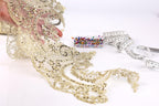 a hand holding a piece of gold lace with intricate patterns, surrounded by various sewing and crafting supplies, including a measuring tape and a small container of colorful buttons.