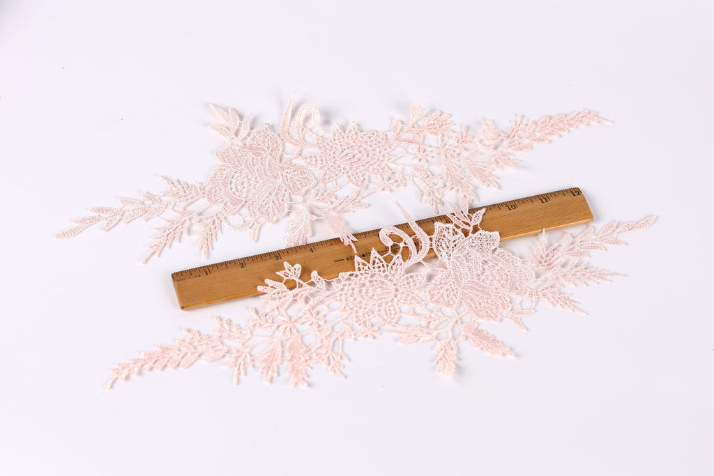 delicate, intricate lace pieces with a floral pattern, arranged on a white background. A wooden ruler is placed in the center of the lace, providing a scale reference for the size of the lace pieces.