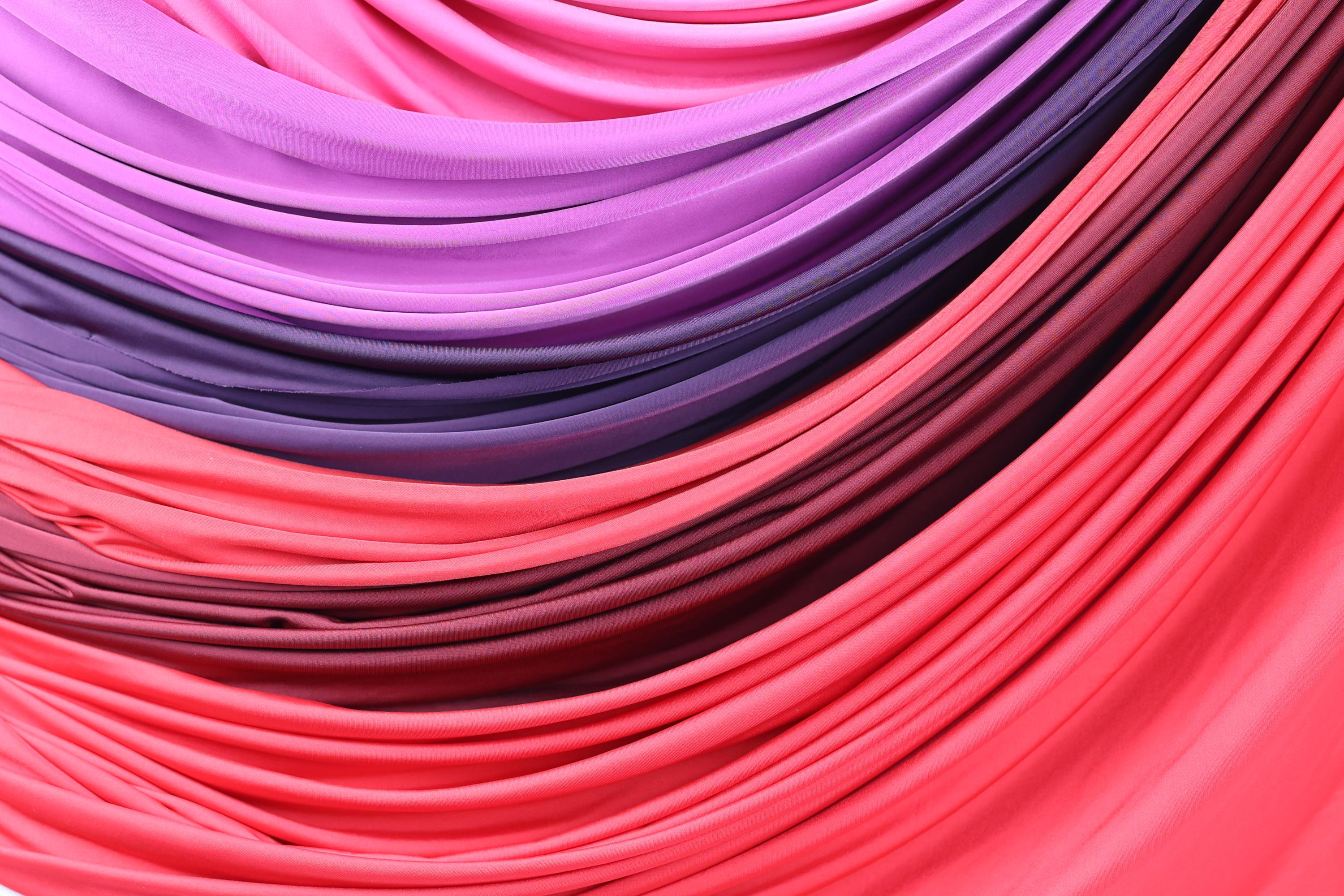 a close-up view of a vibrant fabric with a gradient of colors, transitioning from a deep purple to a bright pink. The fabric appears to be folded or draped in a way that creates a visually striking and dynamic composition.