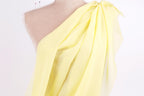 A mannequin wearing a yellow one-shoulder dress with a draped, flowing fabric draped over the shoulder.