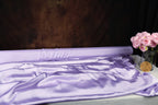 a large roll of purple satin fabric, a vase of pink roses, and a dark brown background.