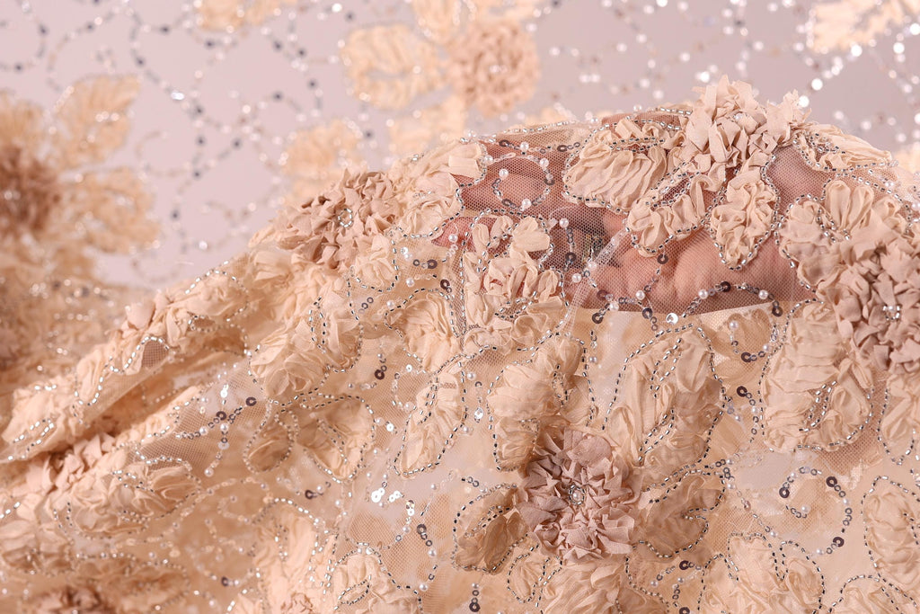 a close-up view of a delicate, intricately embroidered fabric with a floral pattern. The fabric appears to be made of a light-colored material, possibly lace or chiffon, and is adorned with small, sparkling beads or sequins that add a touch of elegance and sparkle to the design. The fabric is folded and draped, showcasing the detailed craftsmanship and artistry involved in its creation.