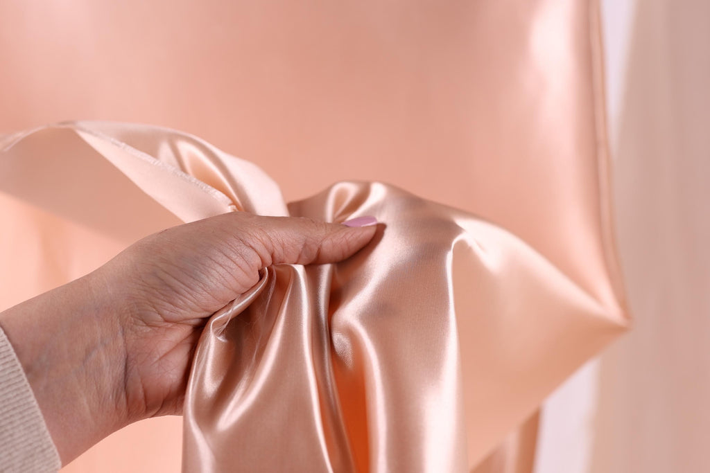 A hand is holding a piece of peach-colored satin fabric, with the fabric partially folded and held in the palm of the hand.