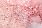 a close-up view of a delicate, pink fabric with a floral pattern. The fabric appears to be made of sheer material, with small sequins or beads scattered throughout the design, adding a touch of sparkle and texture to the overall appearance.