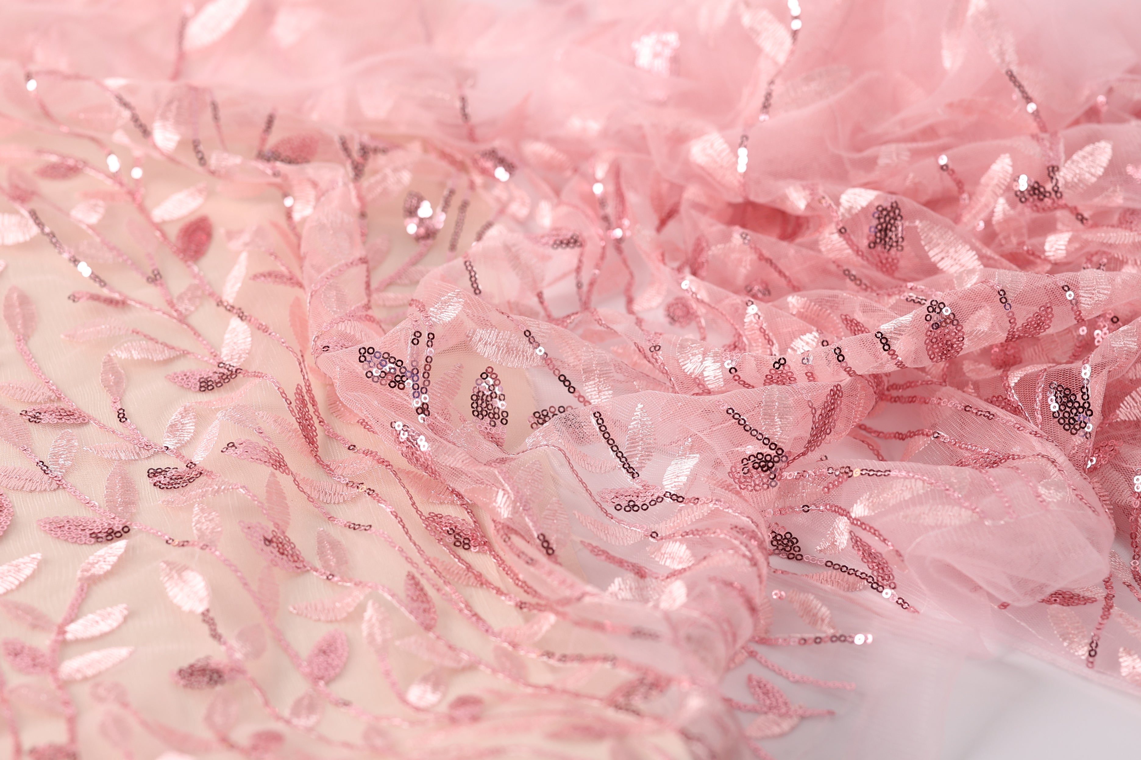 a close-up view of a delicate, pink fabric with a floral pattern. The fabric appears to be made of sheer material, with small sequins or beads scattered throughout the design, adding a touch of sparkle and texture to the overall appearance.