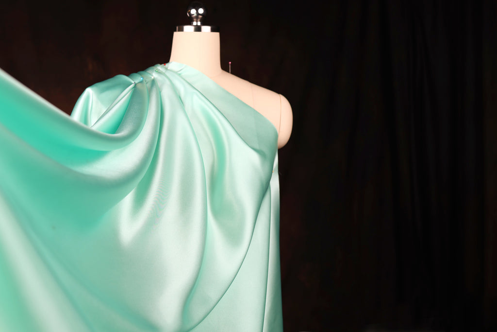 A mannequin wearing a flowing, mint green satin dress with a draped, flowing cape-like design.