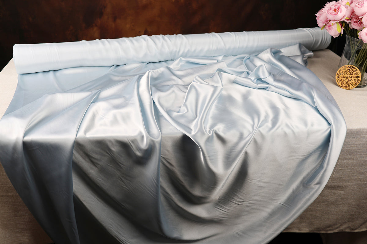 a roll of blue satin fabric on a table, with a vase of pink flowers placed next to it.