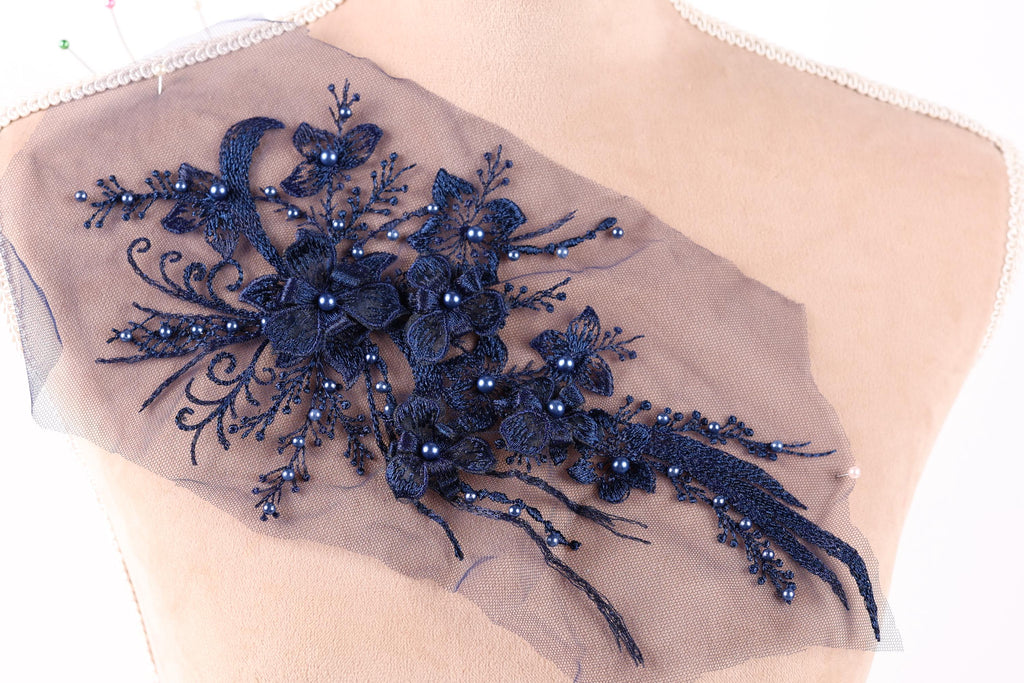 A close-up view of a delicate, intricate blue lace embroidery on a fabric, featuring a floral pattern with blue flowers and pearls.