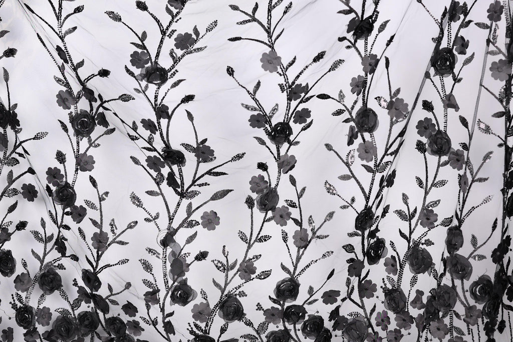 a close-up view of a black and white fabric with a repeating floral pattern. The pattern consists of numerous black flowers and leaves, arranged in a repeating design across the fabric. The fabric appears to be made of a sheer material, allowing the intricate details of the floral pattern to be visible.