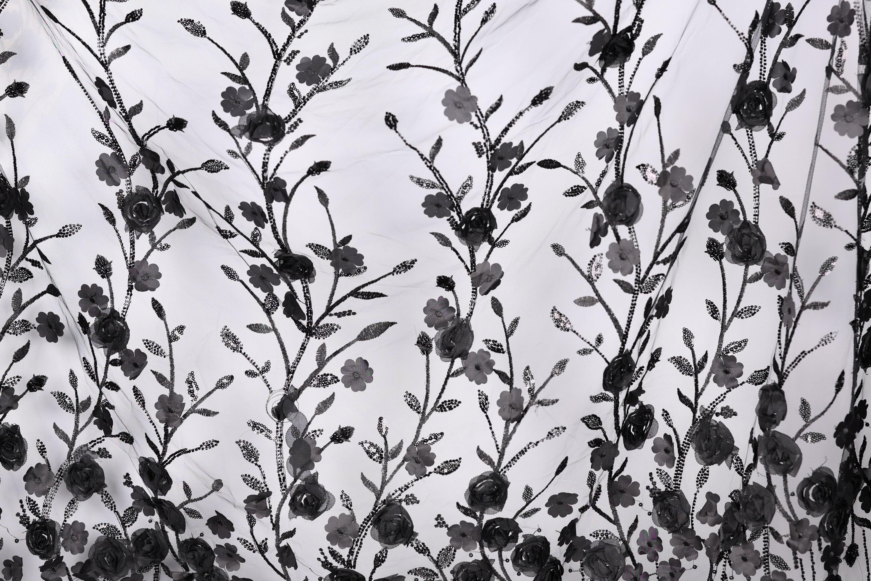 a close-up view of a black and white fabric with a repeating floral pattern. The pattern consists of numerous black flowers and leaves, arranged in a repeating design across the fabric. The fabric appears to be made of a sheer material, allowing the intricate details of the floral pattern to be visible.