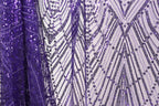a close-up view of a fabric with a purple sequin pattern. The fabric appears to be a sheer material, with a repeating pattern of purple and white sequins. The fabric is displayed in a vertical orientation, with the left side appearing to be slightly higher than the right side. The overall composition of the image is simple and focused on the intricate details of the sequin pattern.