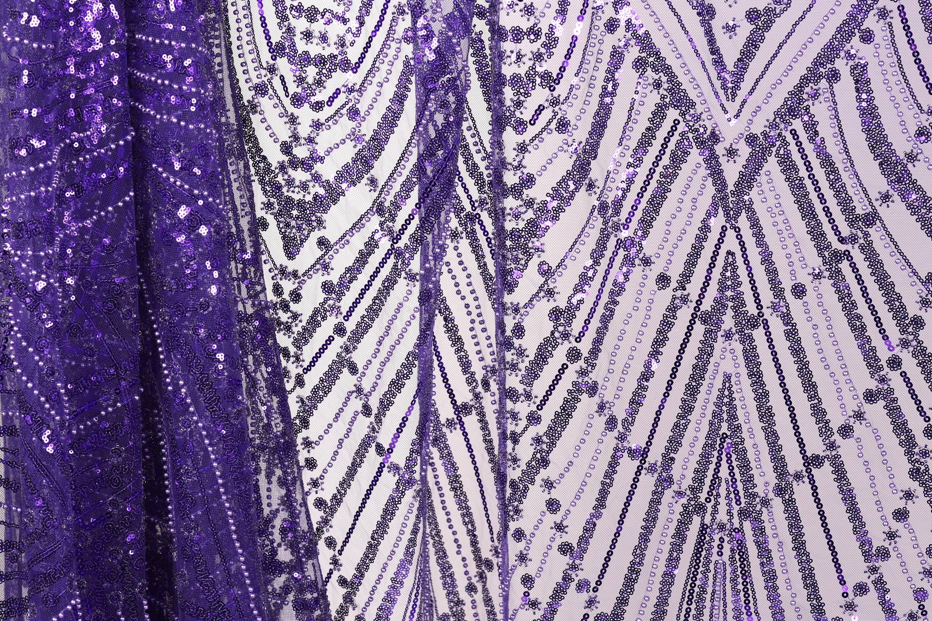 a close-up view of a fabric with a purple sequin pattern. The fabric appears to be a sheer material, with a repeating pattern of purple and white sequins. The fabric is displayed in a vertical orientation, with the left side appearing to be slightly higher than the right side. The overall composition of the image is simple and focused on the intricate details of the sequin pattern.
