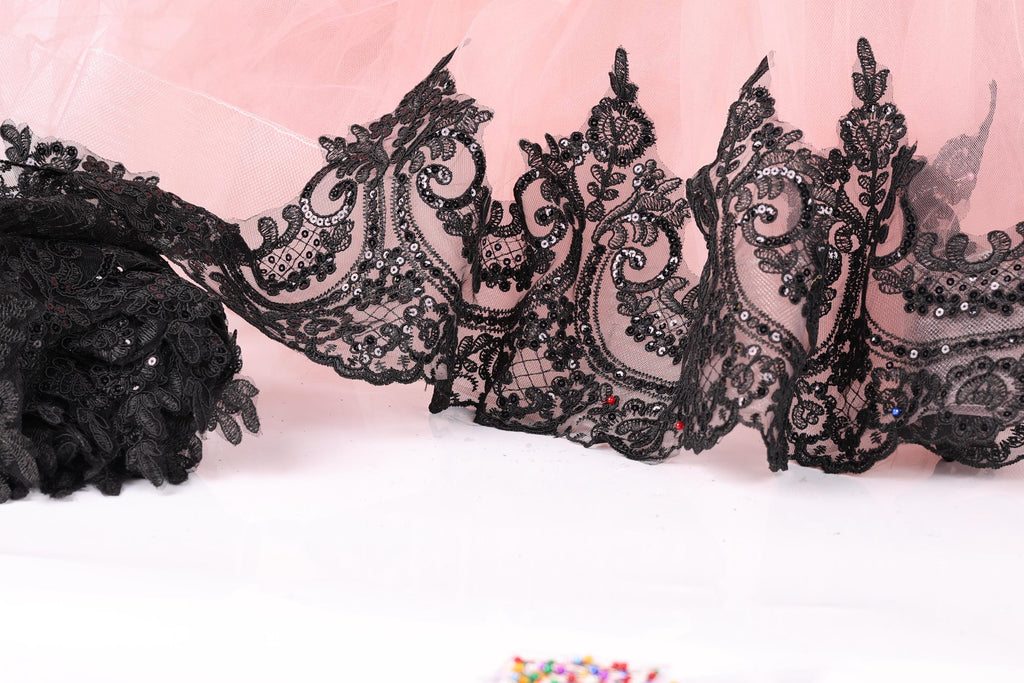 a close-up view of a delicate lace trim with intricate black lace patterns and embellishments, such as sequins or rhinestones, against a pink background.