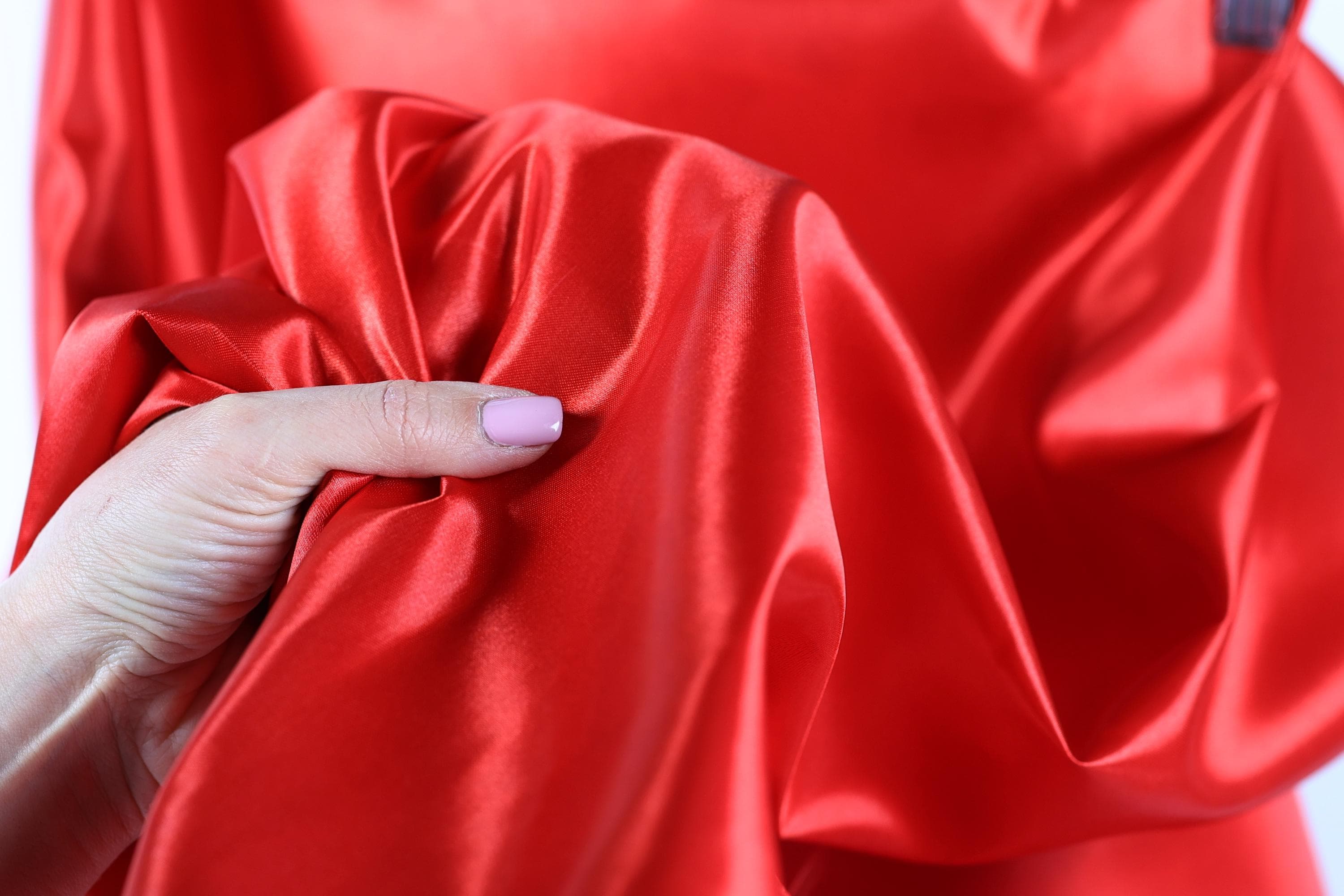A hand is holding a piece of red satin fabric, with the fabric's smooth and shiny texture visible.