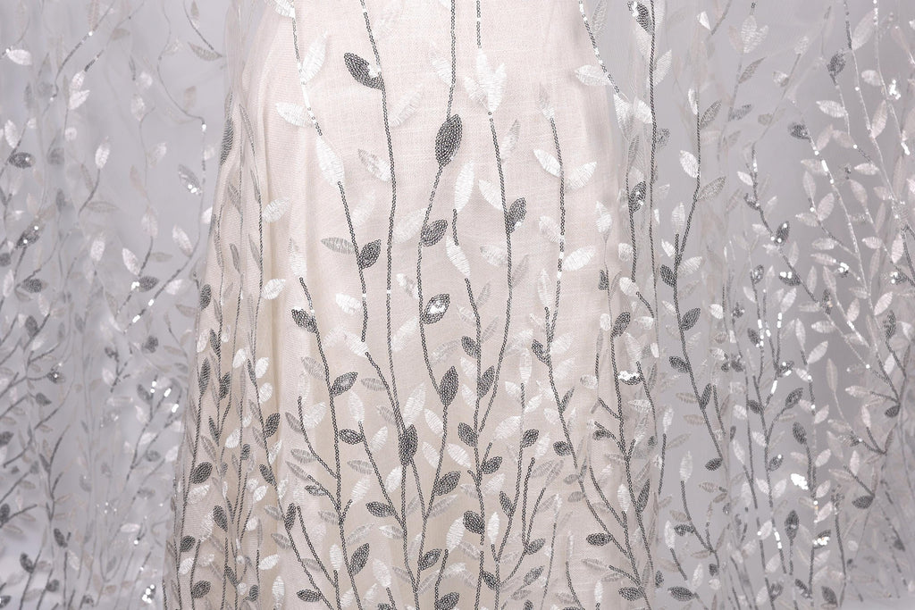 a close-up view of a delicate, white dress with a pattern of silver and white leaves or branches. The dress appears to be made of a sheer fabric, giving it a delicate and elegant appearance.