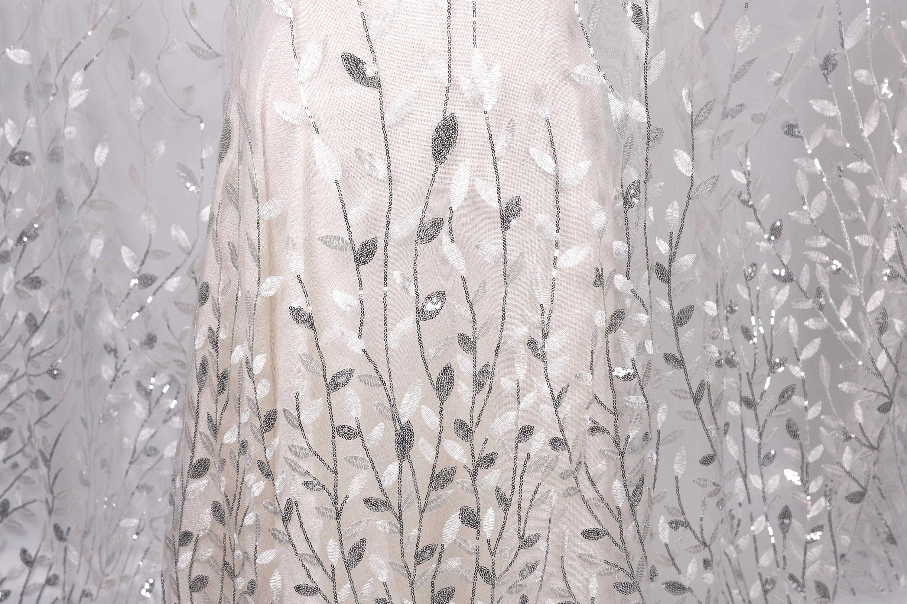 a close-up view of a delicate, white dress with a pattern of silver and white leaves or branches. The dress appears to be made of a sheer fabric, giving it a delicate and elegant appearance.