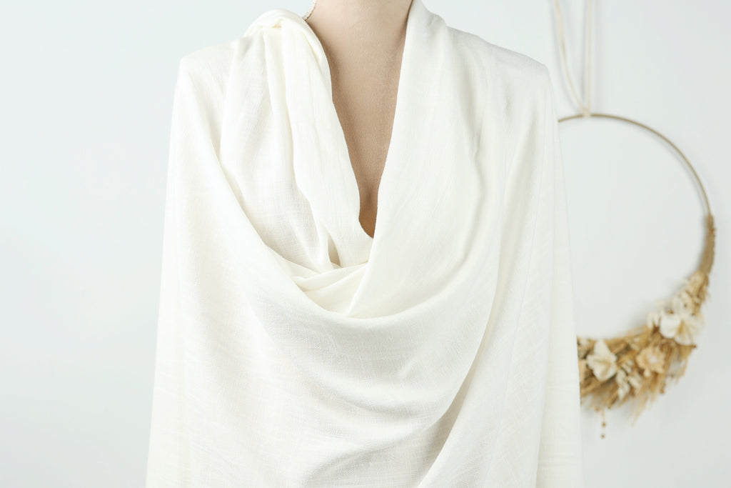 A mannequin wearing a white, draped garment stands in front of a wreath.
