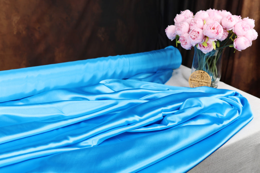 a blue satin fabric draped over a table, with a vase of pink peonies placed next to it.