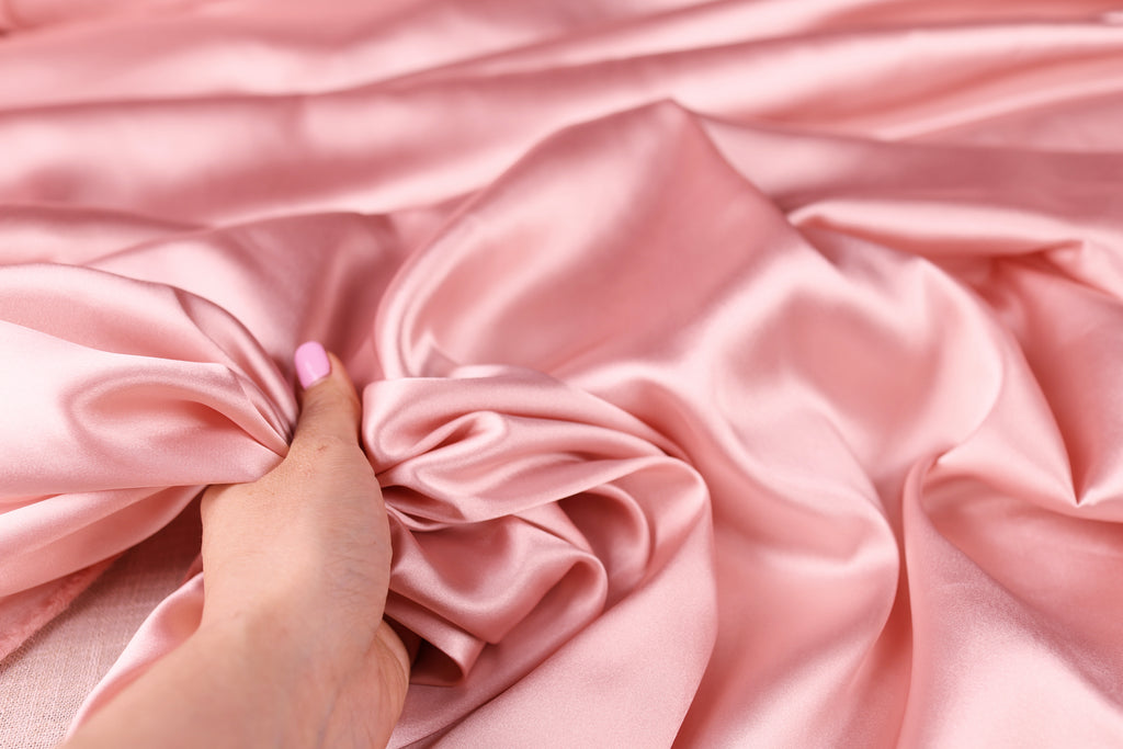 A hand is holding a piece of pink satin fabric, which appears to be soft and smooth.