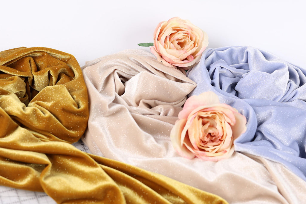 a close-up view of three different fabric textures, including a gold velvet fabric, a beige fabric, and a blue fabric. There are also two pink roses placed on top of the fabrics, adding a touch of color and natural beauty to the composition.