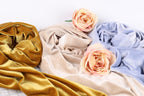 a close-up view of three different fabric textures, including a gold velvet fabric, a beige fabric, and a blue fabric. There are also two pink roses placed on top of the fabrics, adding a touch of color and natural beauty to the composition.