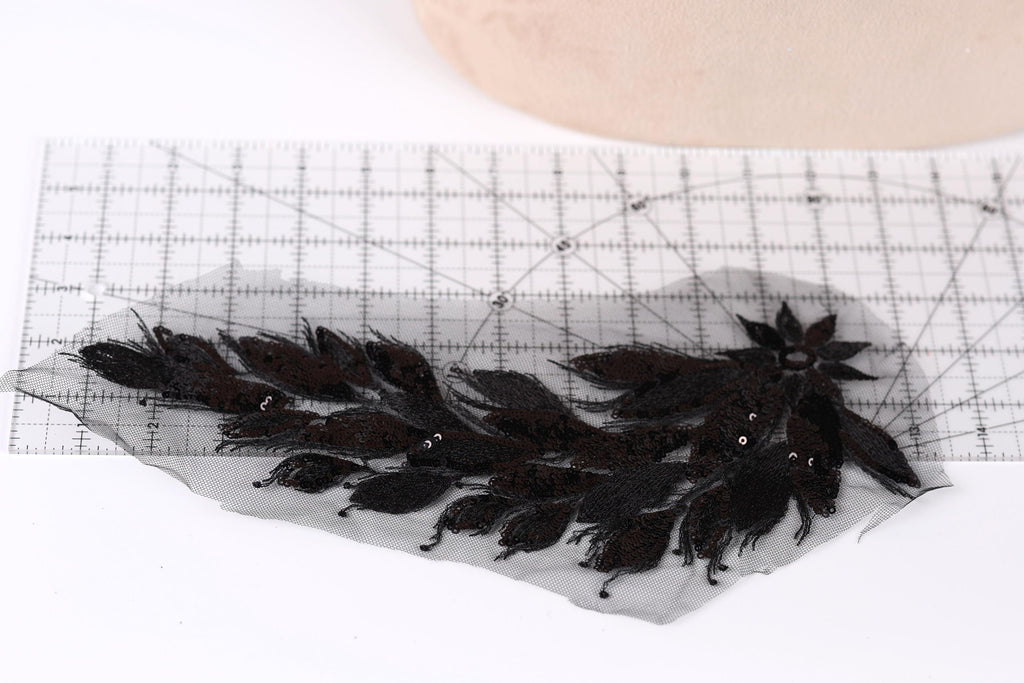 a close-up view of a black lace piece with intricate floral patterns, placed on a ruler for scale.
