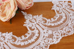a close-up view of a delicate, intricately designed lace fabric with a floral pattern. The lace is placed on a wooden surface, and there is a single pink flower resting on the fabric.