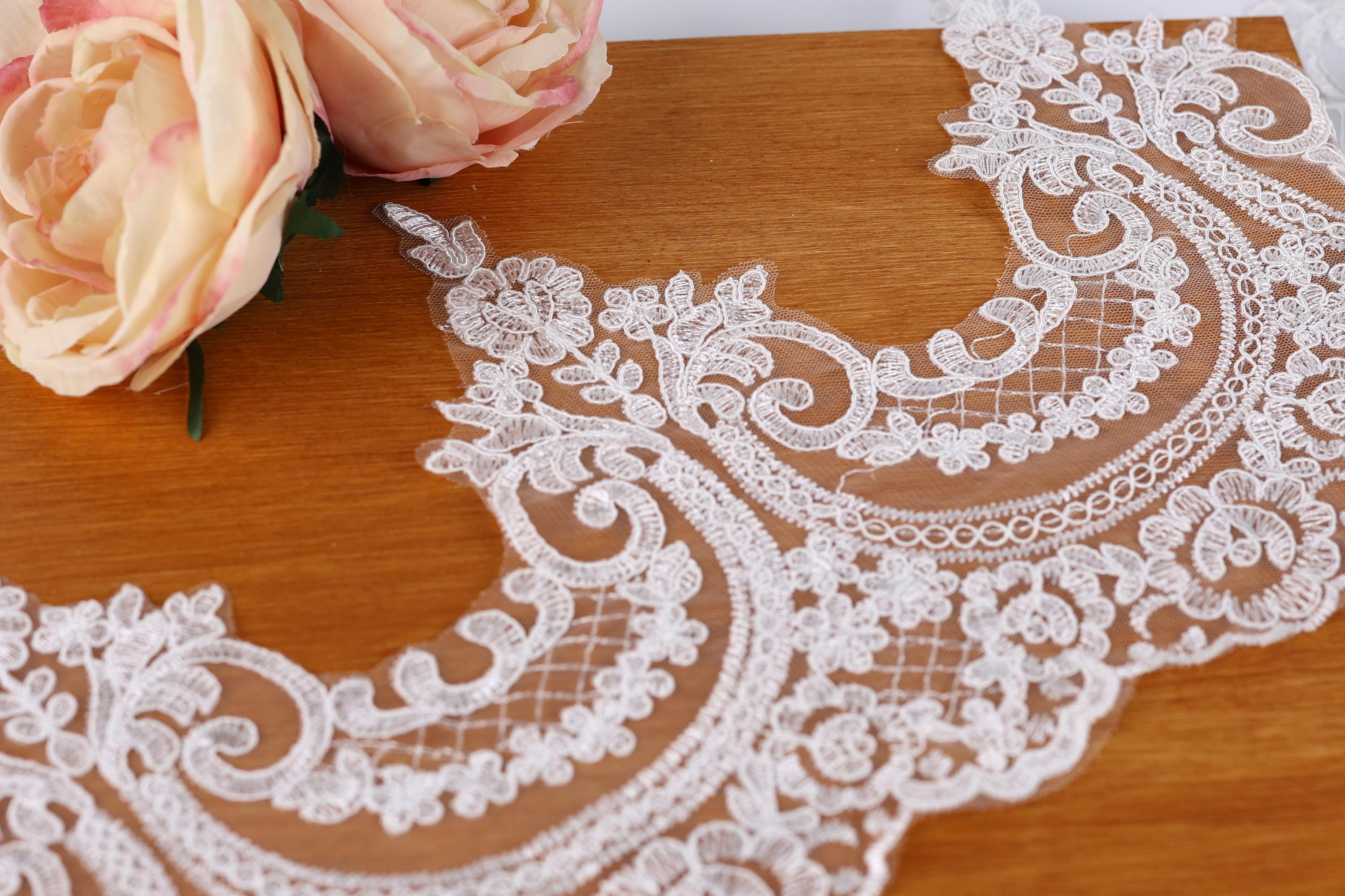 a close-up view of a delicate, intricately designed lace fabric with a floral pattern. The lace is placed on a wooden surface, and there is a single pink flower resting on the fabric.