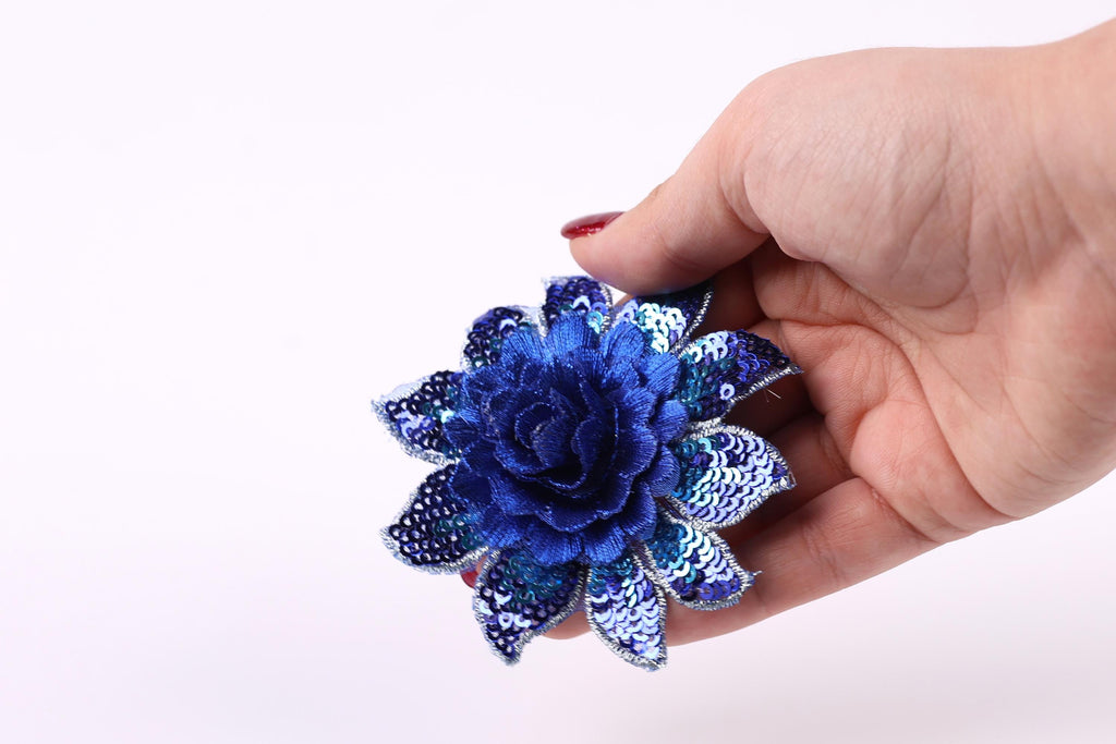 A hand holding a blue sequin flower against a white background.
