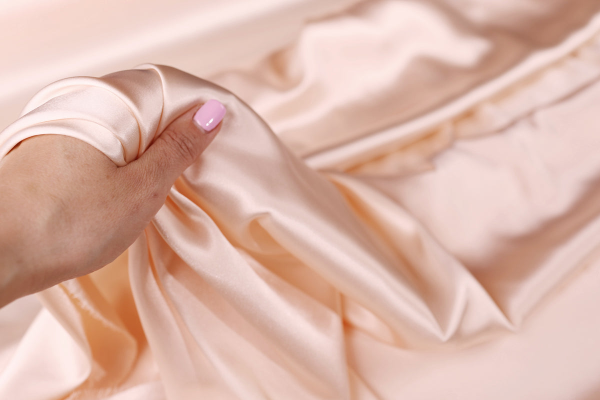 A hand is holding a piece of light pink satin fabric, with the fabric's folds and creases visible.