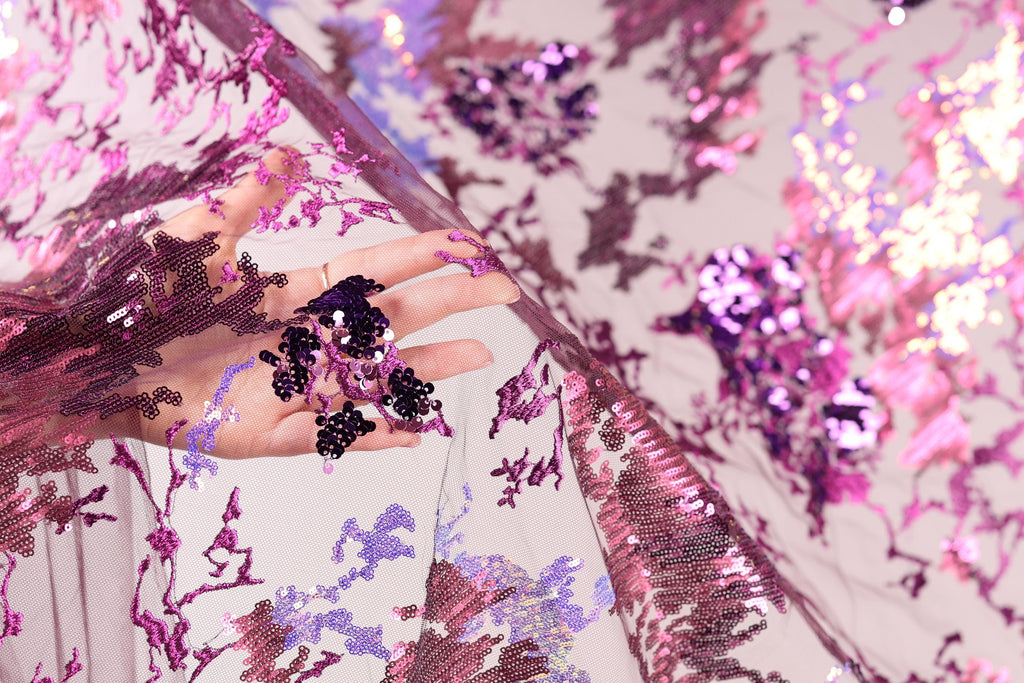 A hand is holding a piece of fabric with a floral pattern of pink and purple sequins.