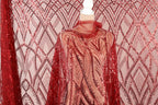 a mannequin wearing a red sequin dress with a sheer overlay. The dress has a pattern of red and white sequins, creating a vibrant and eye-catching design. The mannequin is positioned in front of a pink and white patterned background, which complements the red sequin dress.