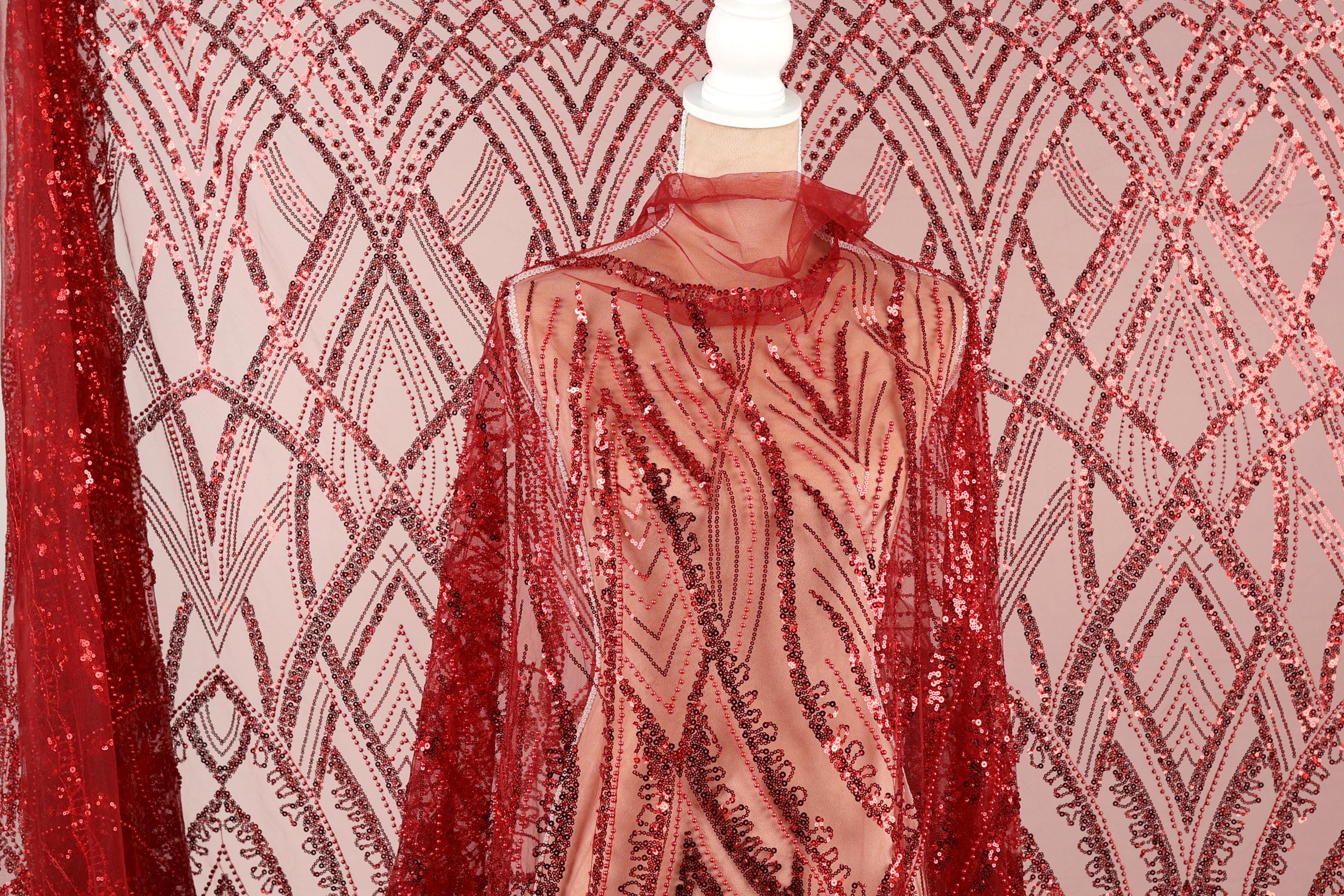 a mannequin wearing a red sequin dress with a sheer overlay. The dress has a pattern of red and white sequins, creating a vibrant and eye-catching design. The mannequin is positioned in front of a pink and white patterned background, which complements the red sequin dress.