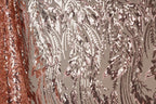 a close-up view of a fabric with a repeating pattern of pink and brown sequin designs. The pattern appears to be a floral or leaf-like design, with the sequins arranged in a way that creates a sense of movement and texture. The fabric is predominantly pink and brown, with the pink sequins standing out against the brown background. The image does not contain any text or other discernible objects, and the focus is solely on the intricate and visually appealing pattern of the sequin fabric.