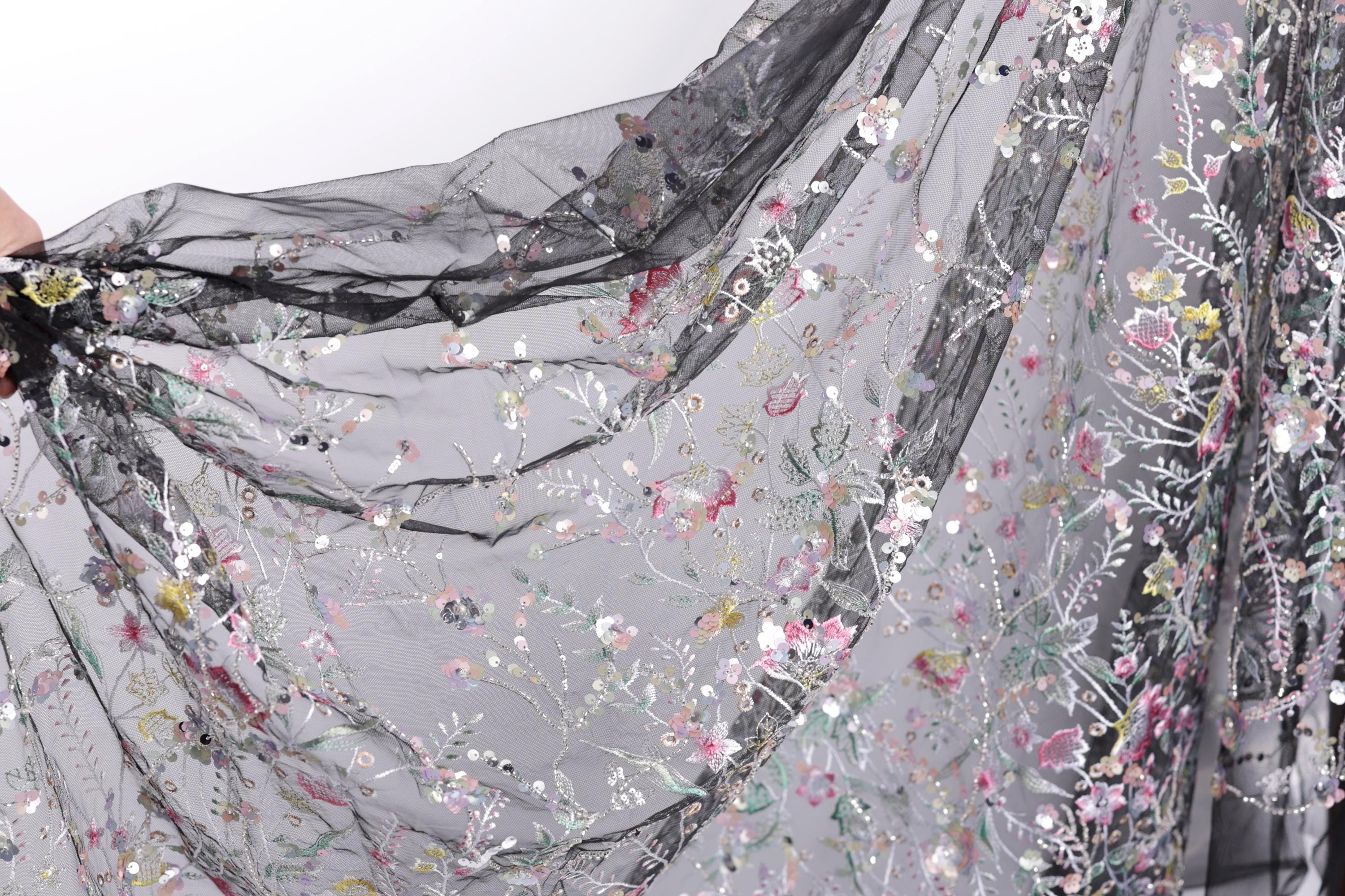 a close-up view of a delicate, intricately embroidered fabric with a floral pattern. The fabric appears to be made of sheer material, giving it a soft and delicate appearance. The embroidery features a variety of flowers and leaves in different colors, creating a visually appealing and detailed design.