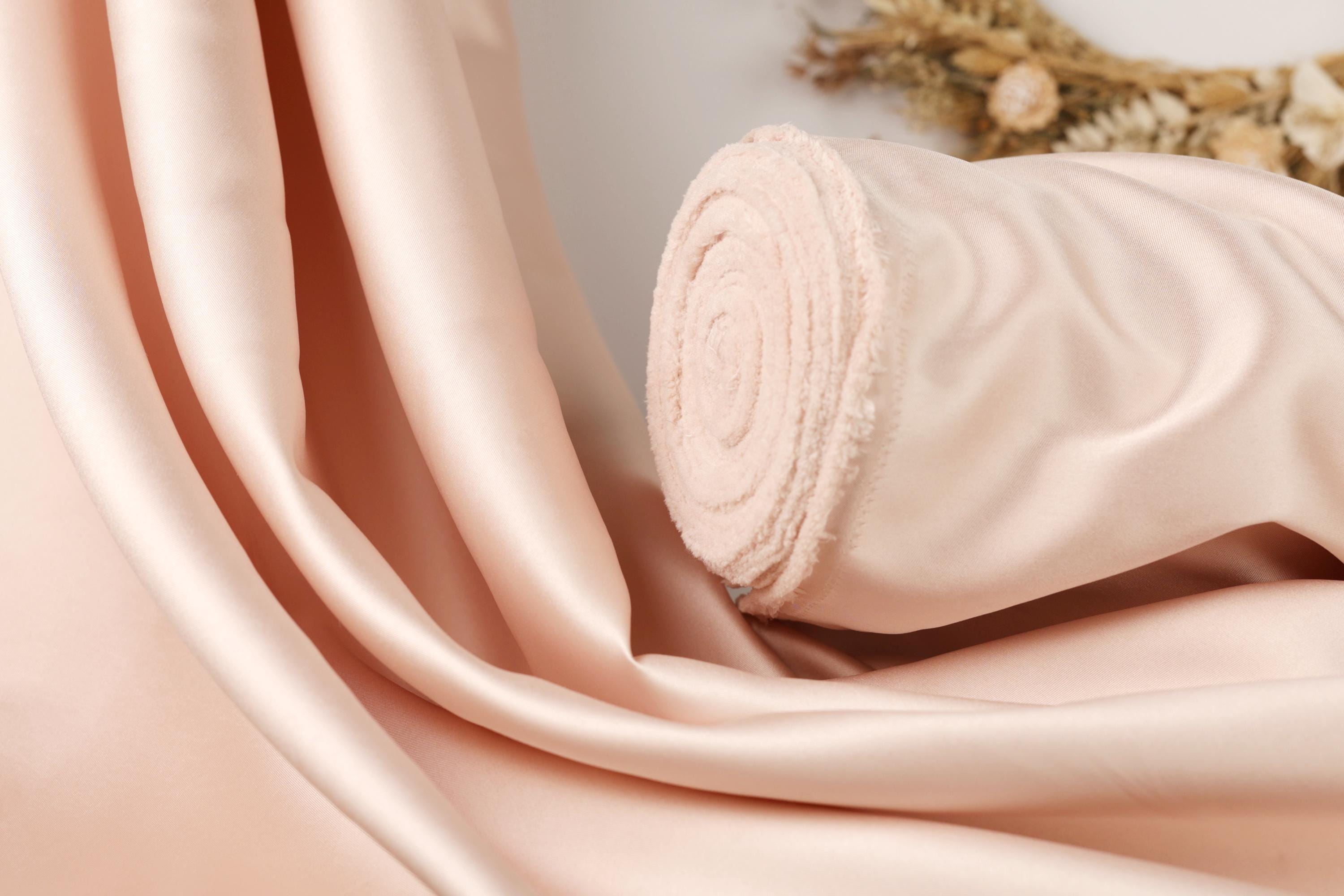 A close-up view of a soft, light pink satin fabric, possibly a sheer or delicate material, with a curled edge and a small amount of fabric visible at the end.