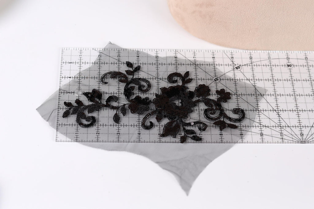 a piece of black lace with intricate floral patterns, placed on a transparent surface. A ruler is visible in the background, indicating that the lace may be used for a sewing or crafting project.