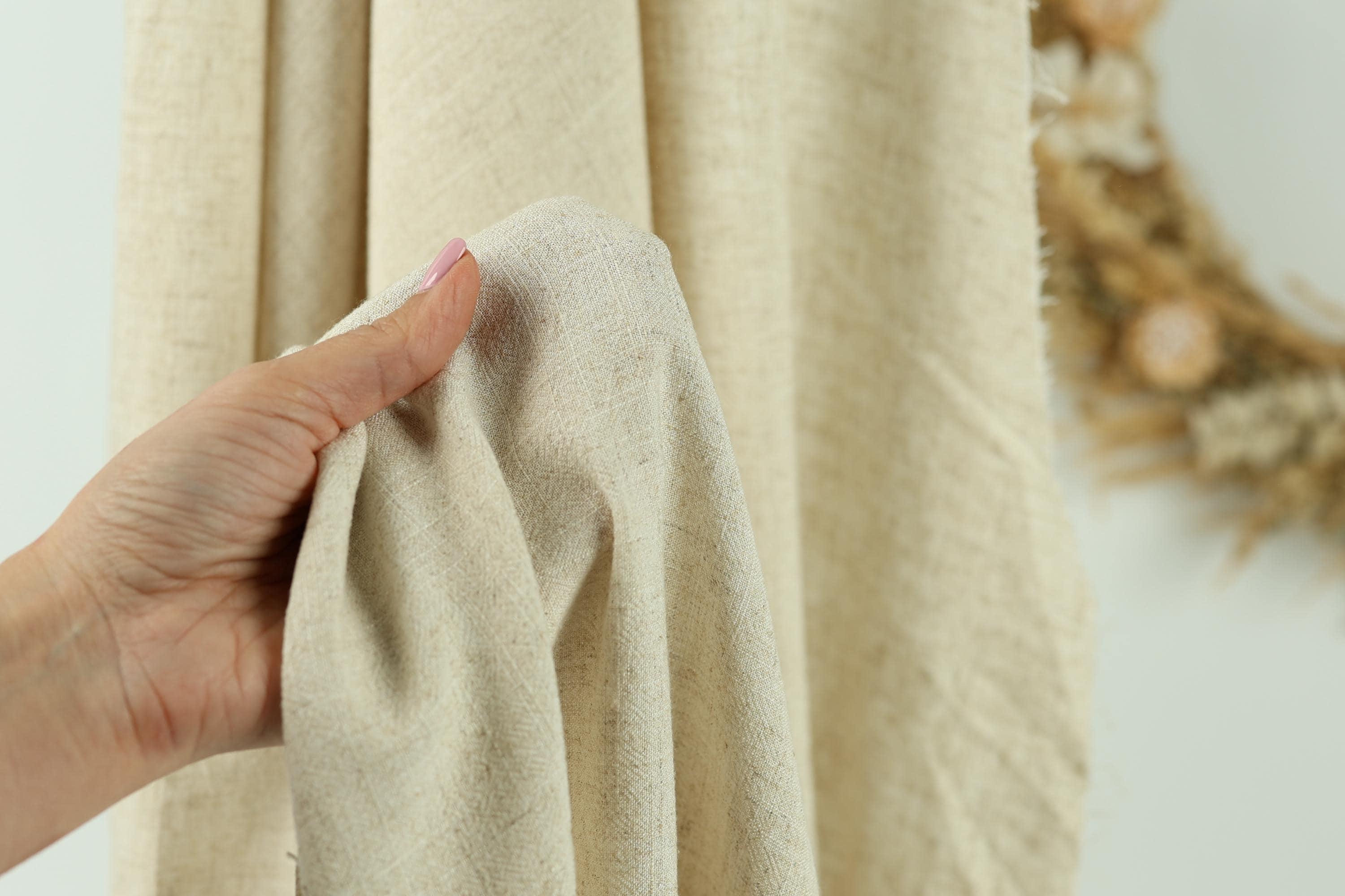 A hand is holding a piece of beige fabric, possibly a curtain or textile, in front of a blurred background.
