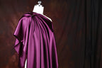 A mannequin wearing a purple satin dress with a draped, flowing silhouette against a dark background.