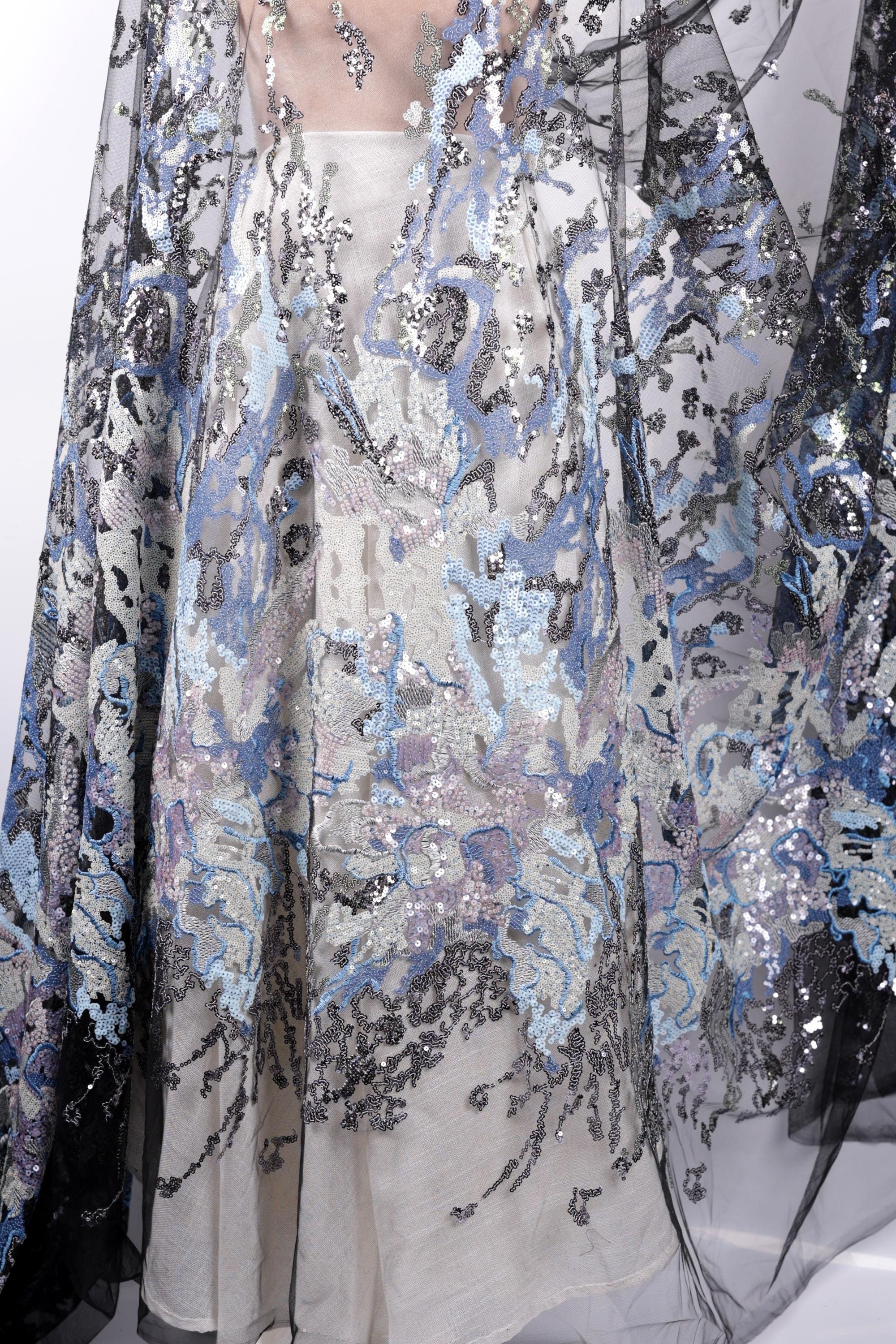 a close-up view of a fabric with a floral pattern and sequin details. The fabric appears to be a sheer material, with a mix of blue, gray, and black colors. The pattern is intricate and detailed, showcasing the craftsmanship of the fabric.