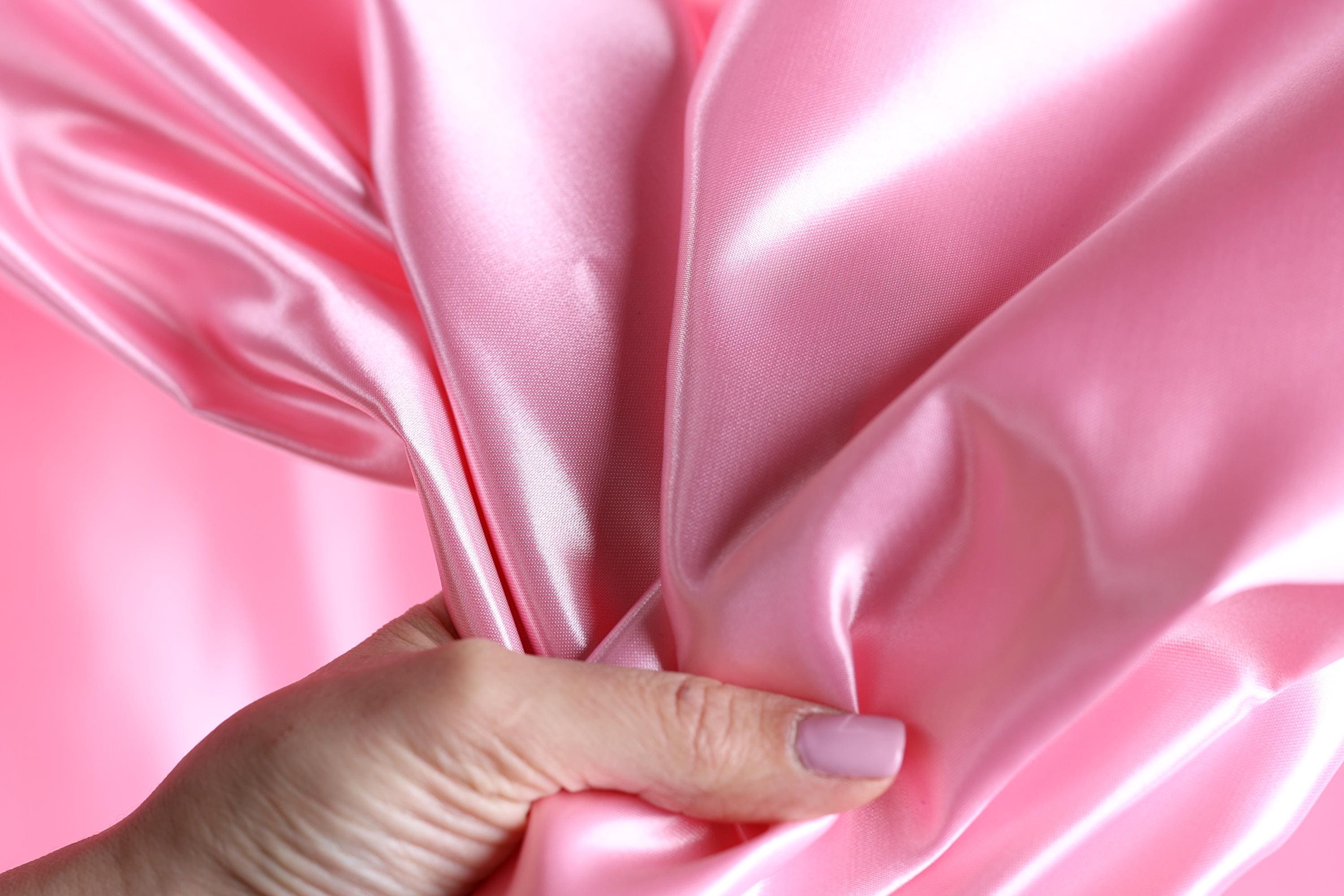 A hand is holding a piece of pink satin fabric, with the fabric partially draped over the hand.