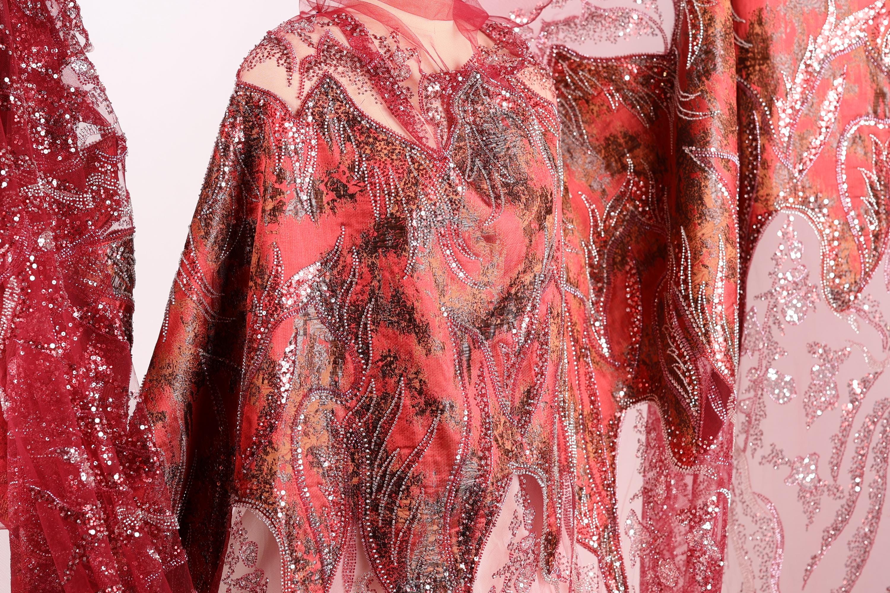 a close-up view of a vibrant red and pink fabric with intricate patterns and designs, including a floral motif and sparkling embellishments.