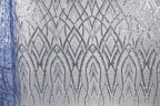 a close-up view of a fabric with a repeating pattern of silver and blue sequin designs. The pattern consists of a series of arched shapes and lines that create a visually striking and intricate design. The fabric appears to be a type of sheer material, with the silver and blue sequins adding a touch of sparkle and elegance to the overall appearance.