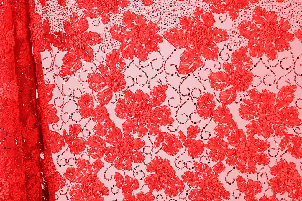 a close-up view of a fabric with a floral pattern in shades of red and pink. The fabric appears to be made of a sheer material, with a repeating pattern of red flowers and leaves. The background of the image is a light pink color, which contrasts with the red of the fabric. The image does not contain any text or other discernible objects or actions.