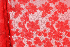a close-up view of a fabric with a floral pattern in shades of red and pink. The fabric appears to be made of a sheer material, with a repeating pattern of red flowers and leaves. The background of the image is a light pink color, which contrasts with the red of the fabric. The image does not contain any text or other discernible objects or actions.