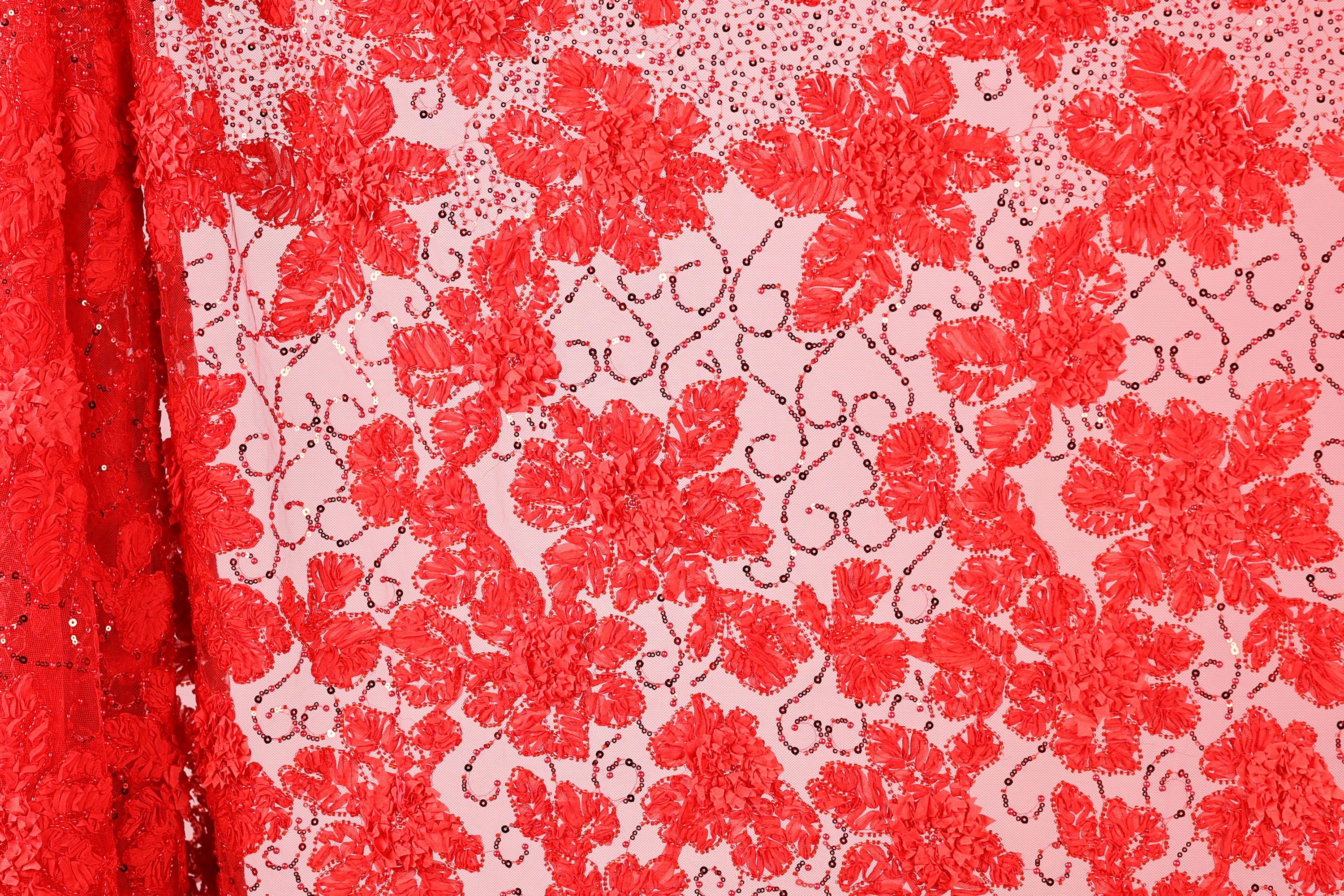a close-up view of a fabric with a floral pattern in shades of red and pink. The fabric appears to be made of a sheer material, with a repeating pattern of red flowers and leaves. The background of the image is a light pink color, which contrasts with the red of the fabric. The image does not contain any text or other discernible objects or actions.