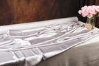 a white satin fabric draped over a table, with a vase of pink flowers placed next to it.