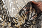 A close-up view of a hand holding a piece of sequin fabric, with the fabric's intricate patterns and shimmering gold and black sequins clearly visible.